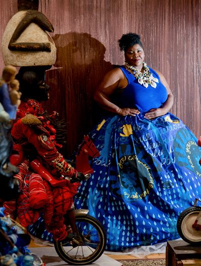 Person in vibrant blue dress in artistic space with sculptures and wooden surfaces.