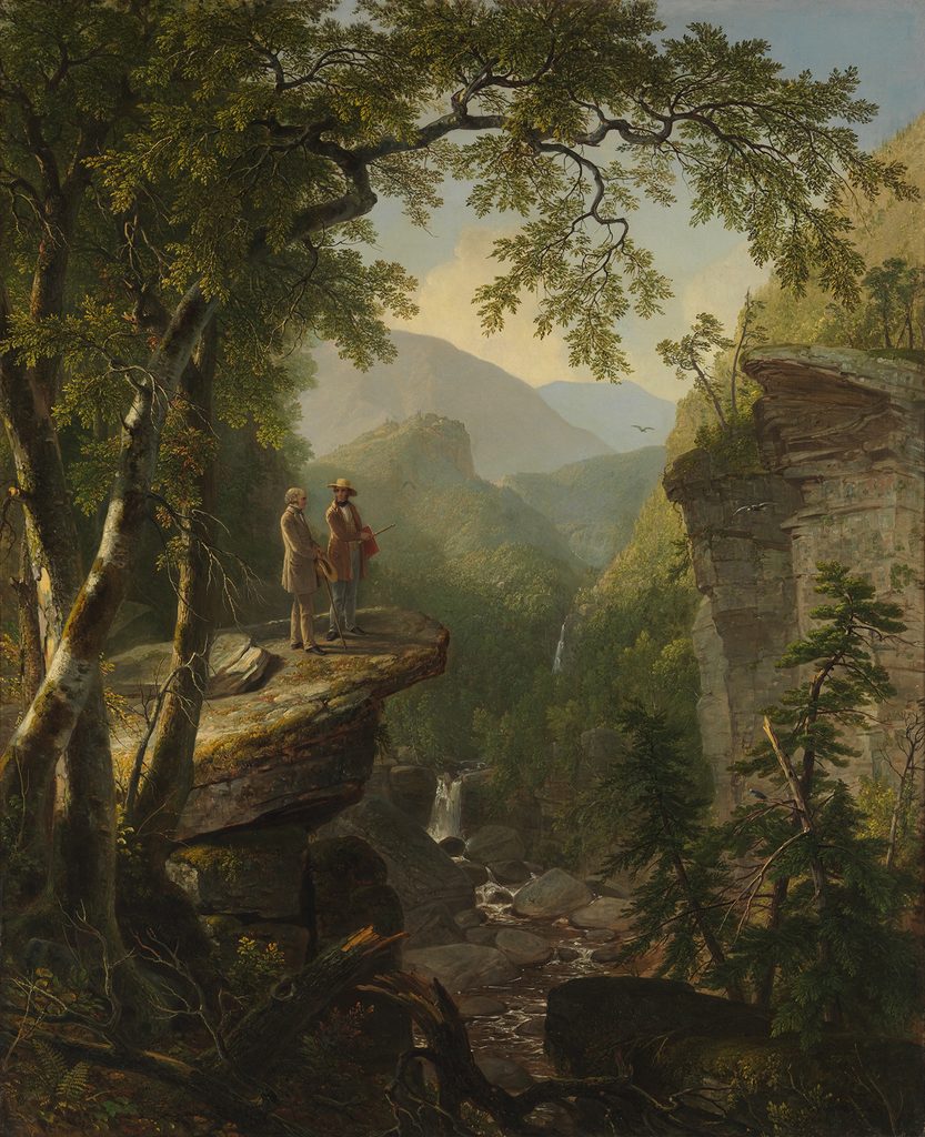Two people standing on a rocky cliff overlooking a forested valley with waterfall and mountains.