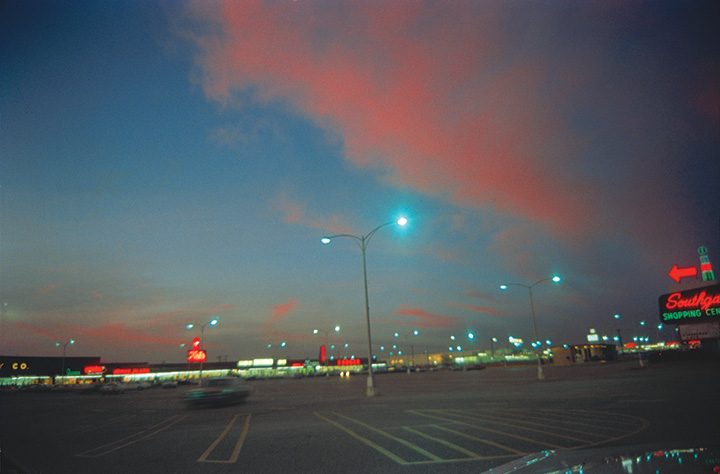 Shopping center parking lot at dusk with streetlights and pink-blue sky, store signs in view.