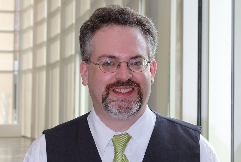 Man with glasses, short hair, goatee; wearing white shirt, dark vest, green tie, bright interior.
