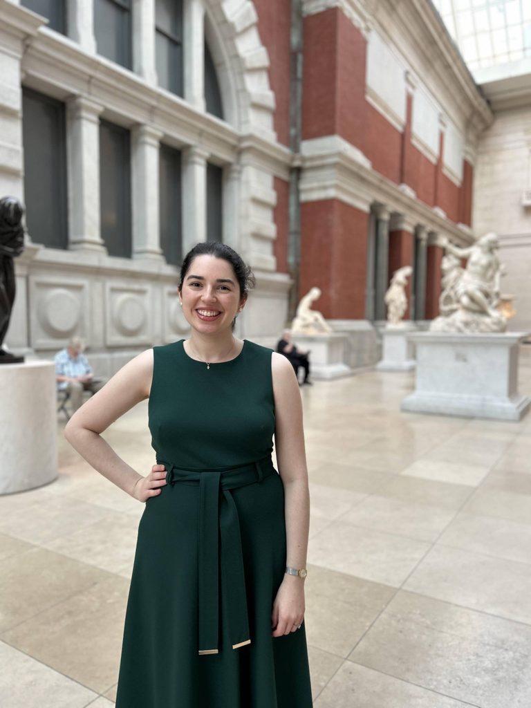 Person in dark green dress stands in a museum with classical sculptures and large windows.