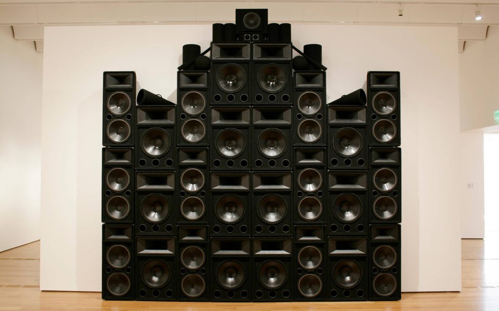 A large structure of stacked black speakers, black megaphones and black cylinders towers next to a white wall