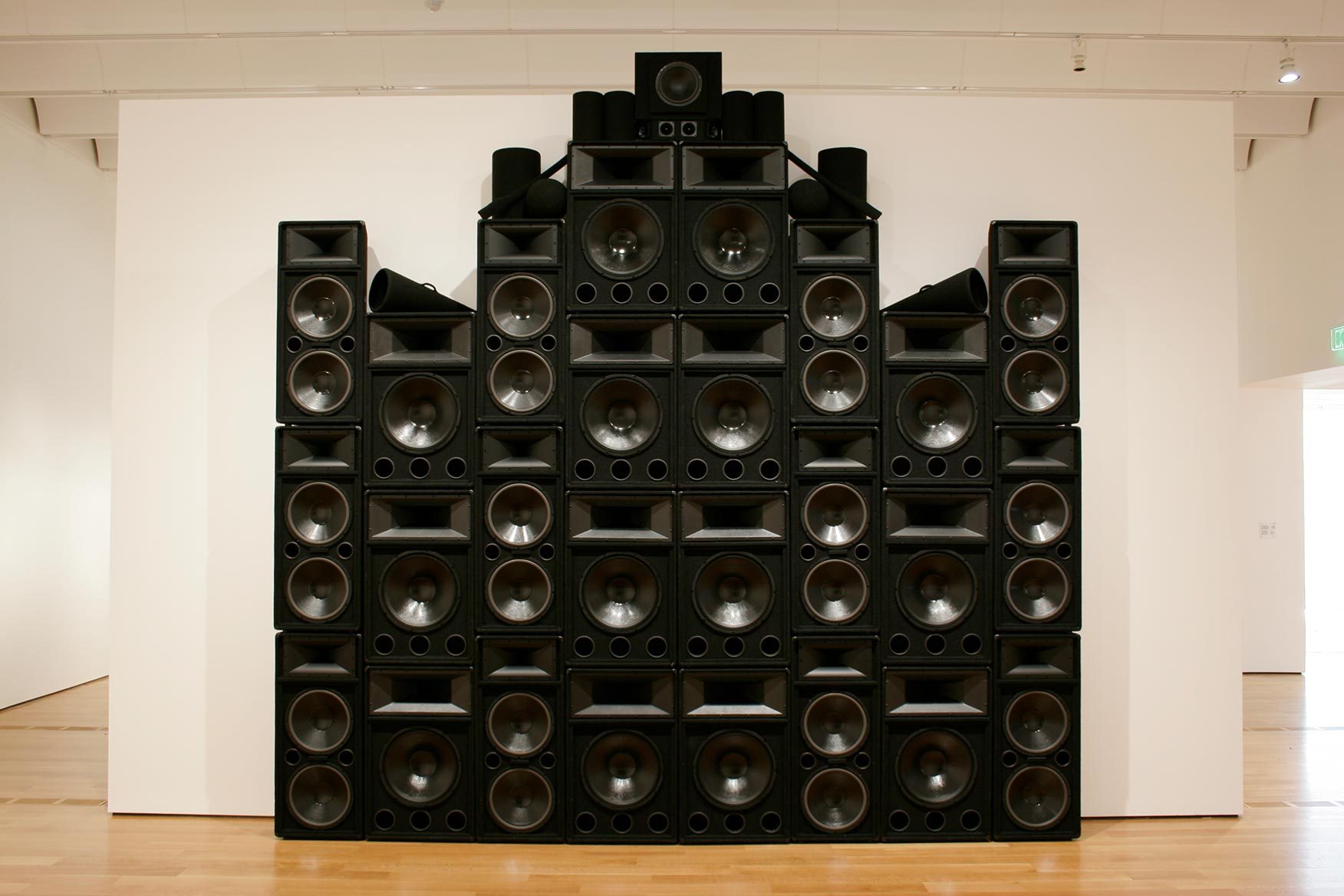 A large structure of stacked black speakers, black megaphones and black cylinders towers next to a white wall