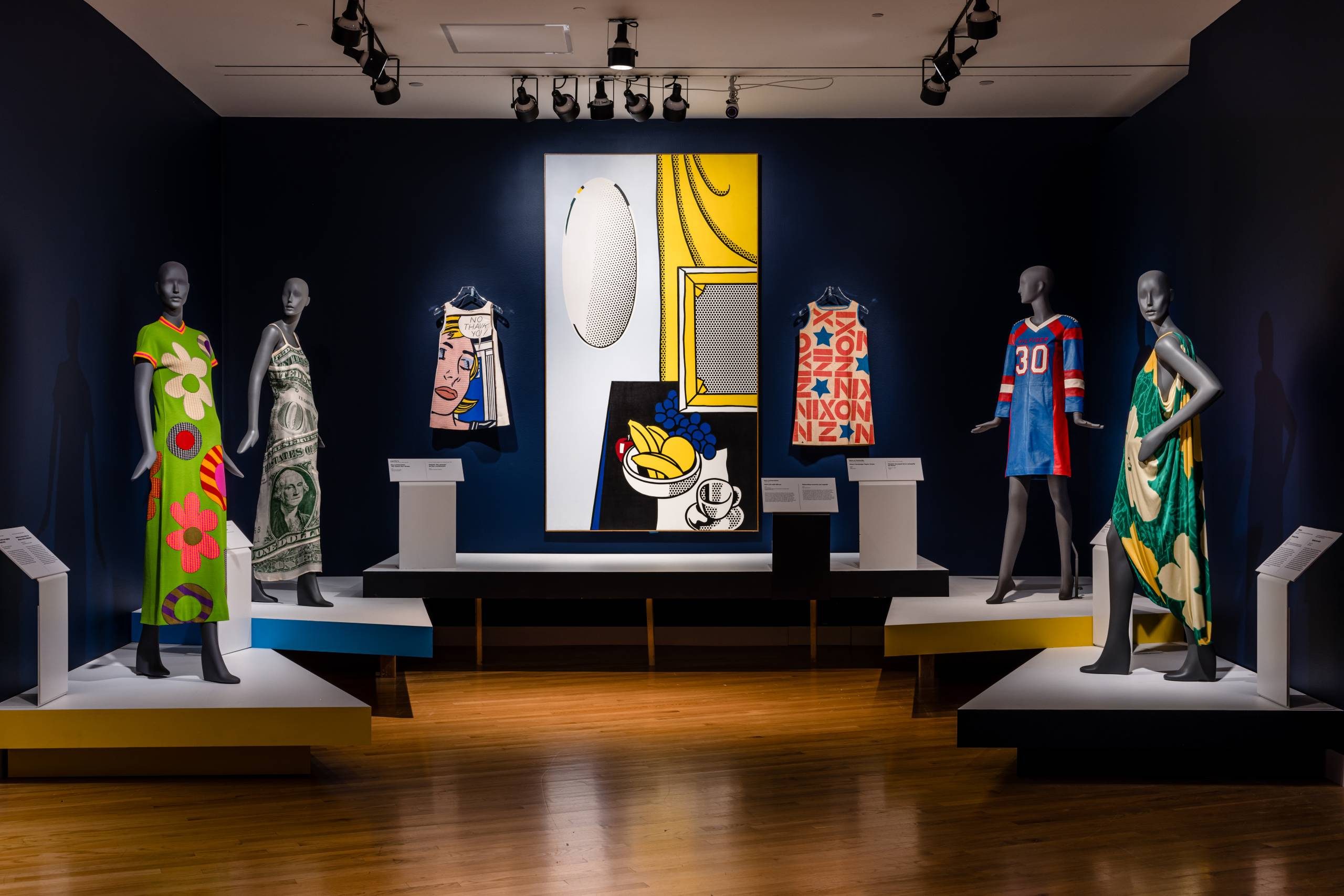 a gallery space featuring mannequins dressed in pop art-inspired garments with bright colors and patterns framing Roy Lichtenstein's work 