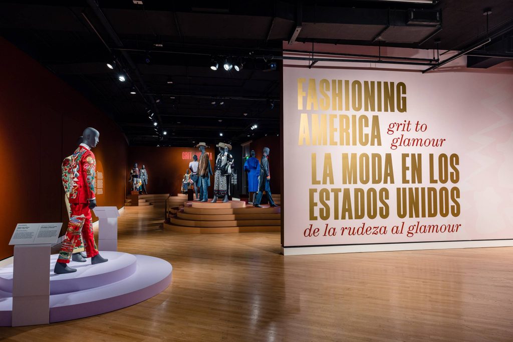 a gallery space featuring mannequins dressed in nudie suits and a bright pink wall featuring the exhibition's title, 