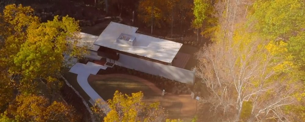 Modern flat-roofed house in autumn trees, white walkway, landscaped areas, aerial view.