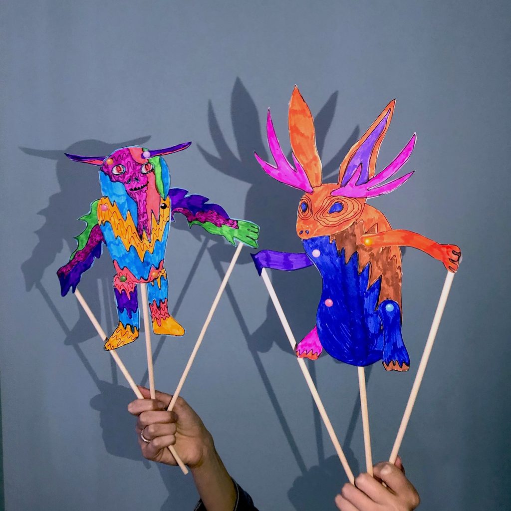 Two colorful paper puppets on sticks, with fantasy designs, casting shadows on a plain background.