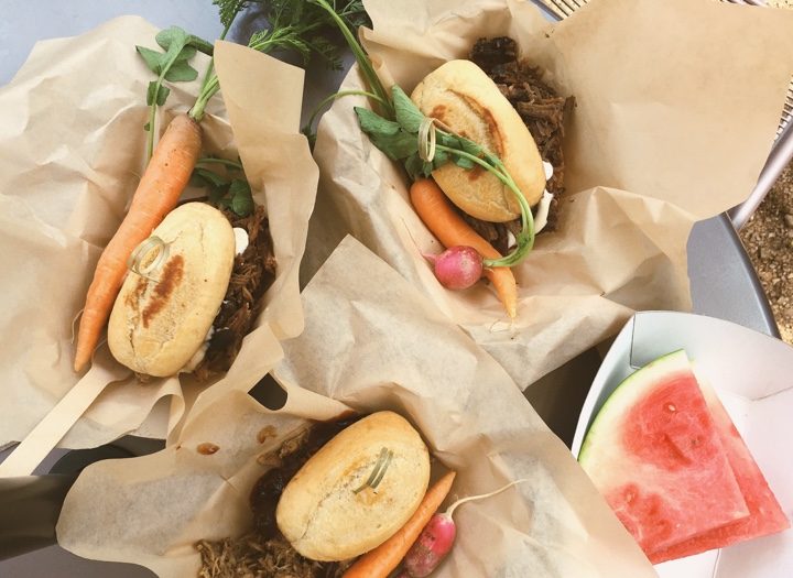 Three brown-paper-wrapped sandwiches with carrot, radish, and nearby watermelon slice on a plate.