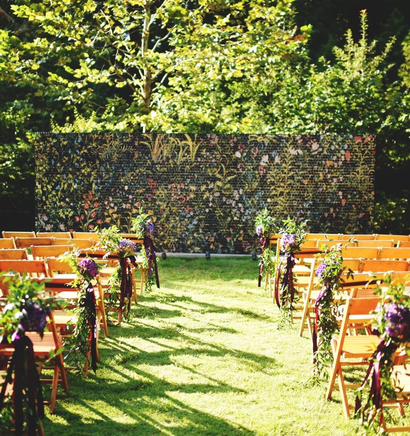 Outdoor event setup with decorated chairs, floral wall, and trees in the background.