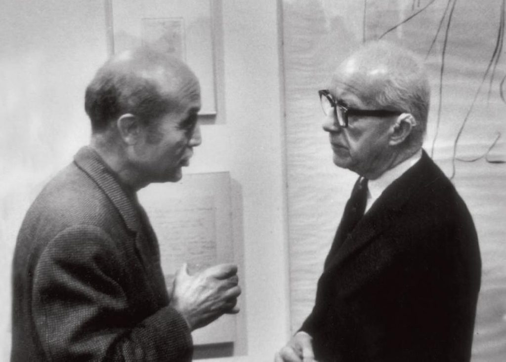 Two men conversing in a gallery with framed art; one holds an object, the other wears glasses.