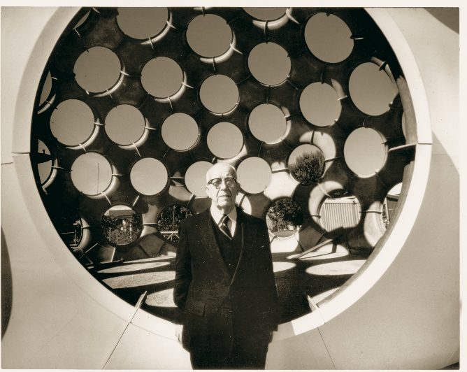 Elderly man in formal attire before a structure with circular openings in sepia tones.