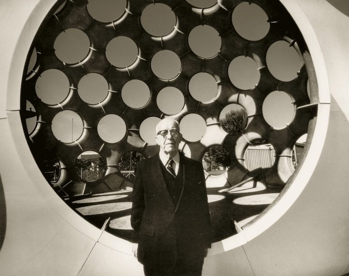 Older man in suit stands before circular structure with circles, trees and fence in background.