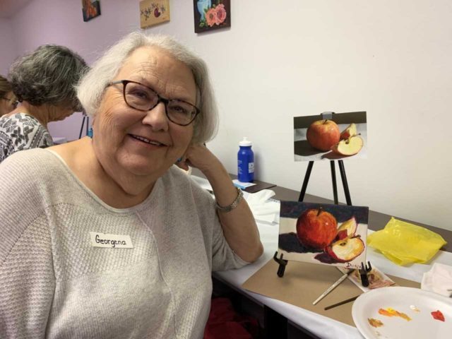 Elderly woman smiles at a table with painting of apples, art supplies, and reference photo.