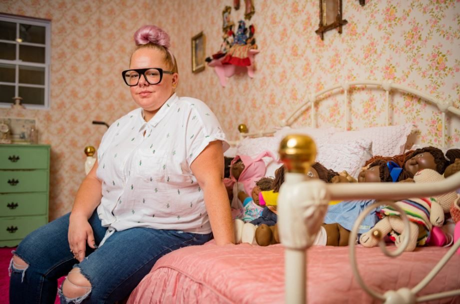 Person with topknot and glasses on vintage bed with dolls, pink bedspread, floral wallpaper.