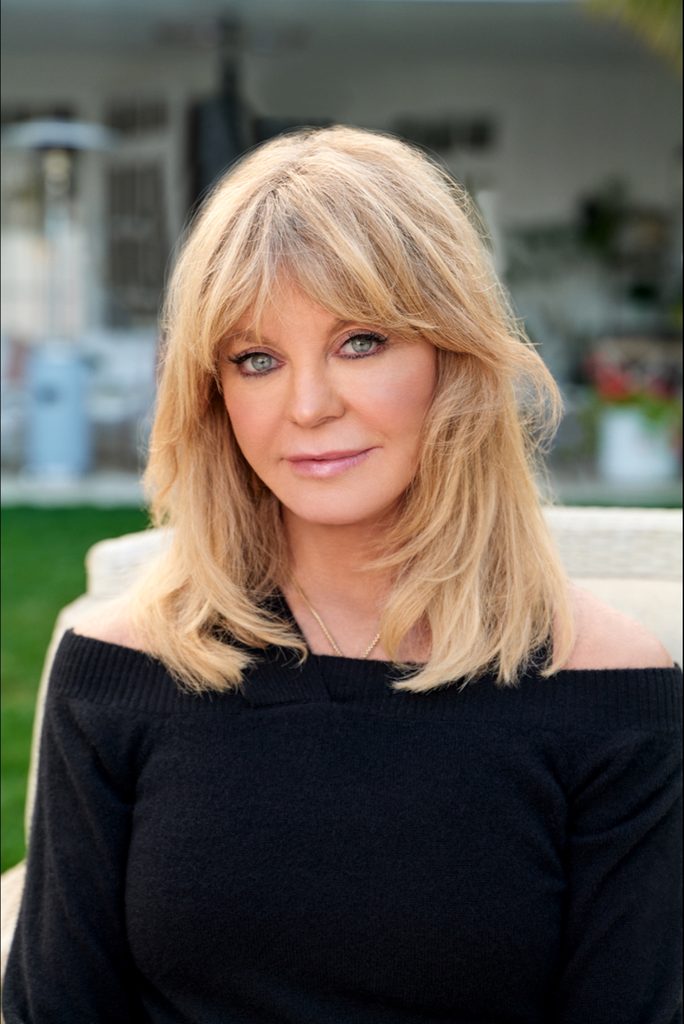 Blonde woman wearing a black off-shoulder top and necklace sitting outdoors with blurred background.
