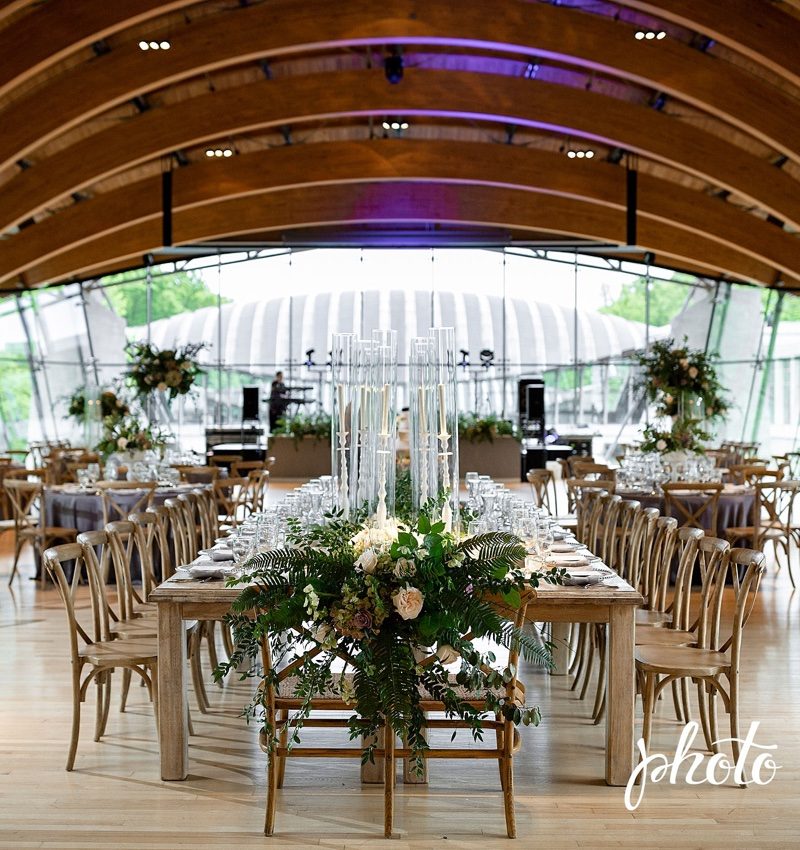 Elegant dining venue with wooden tables, floral decor, glass candles, arched ceiling, large windows.