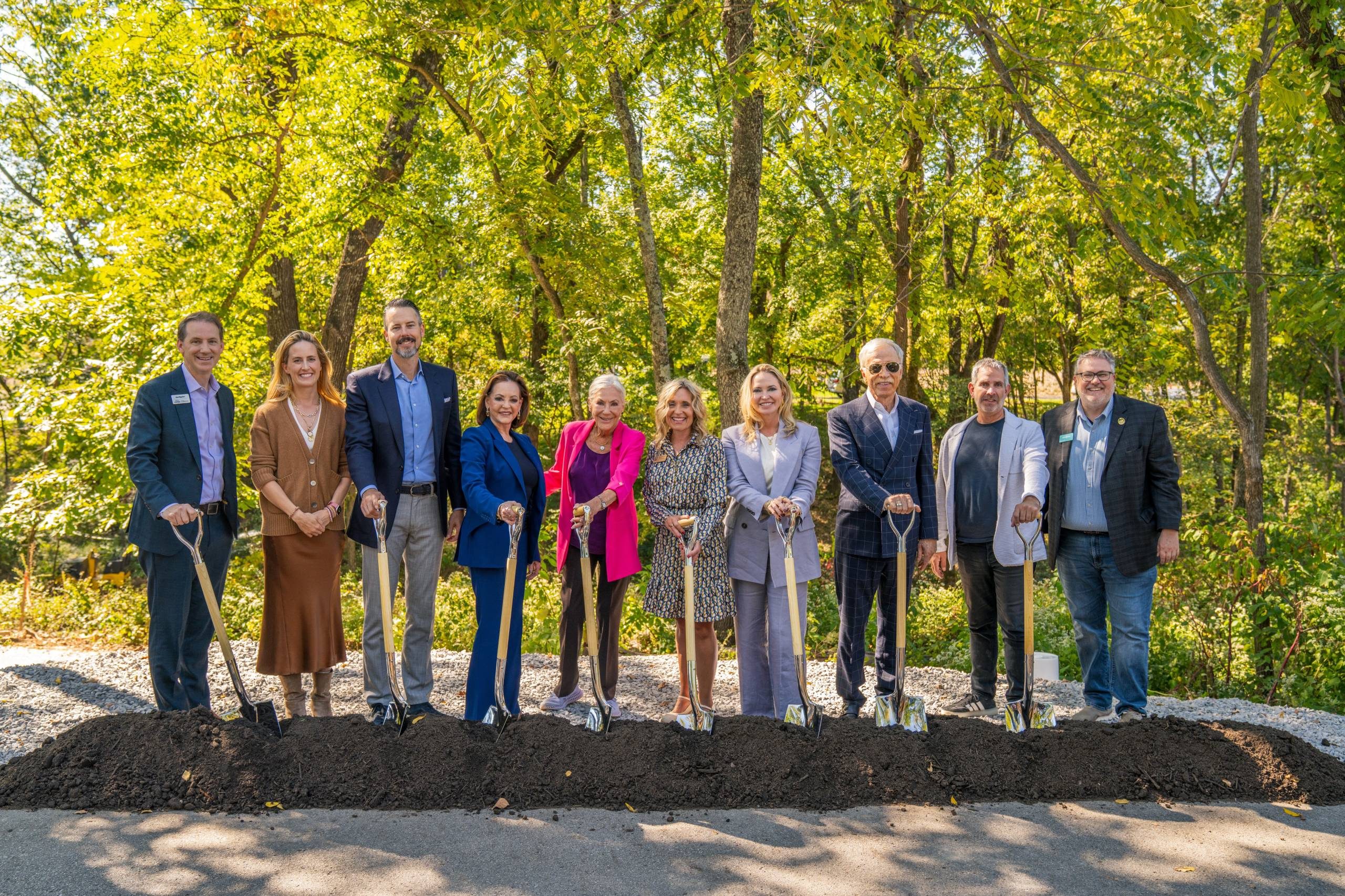 Group of ten people holding shovels at a groundbreaking ceremony outdoors with trees behind.