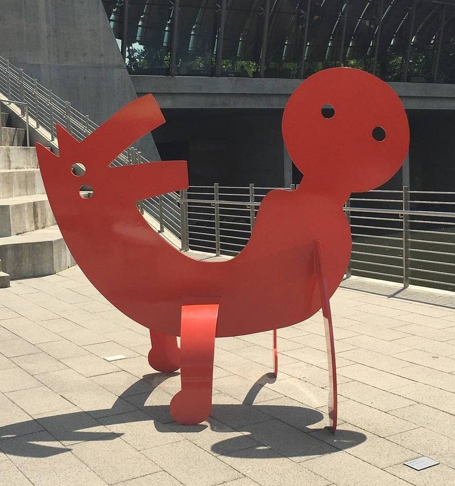 Red abstract metal sculpture of reclining figure with round head and circular eyes on paved area.