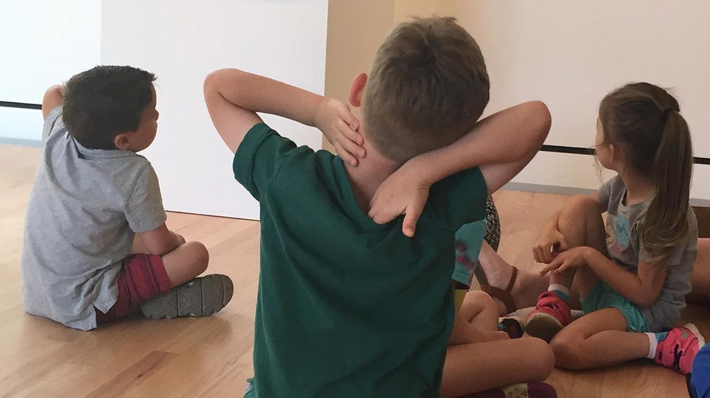 Children sitting on a wooden floor in an informal setting, one with hands on head.