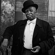 Older man in bowler hat and suit with bow tie standing by brick wall and railing.