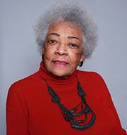 Elderly woman with gray hair, red turtleneck, black necklace, plain background.