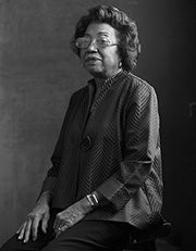 Elderly woman seated, wearing glasses and patterned top, in a monochromatic setting.