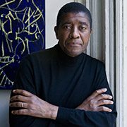 Person in black turtleneck with crossed arms, standing beside abstract artwork on white wall.