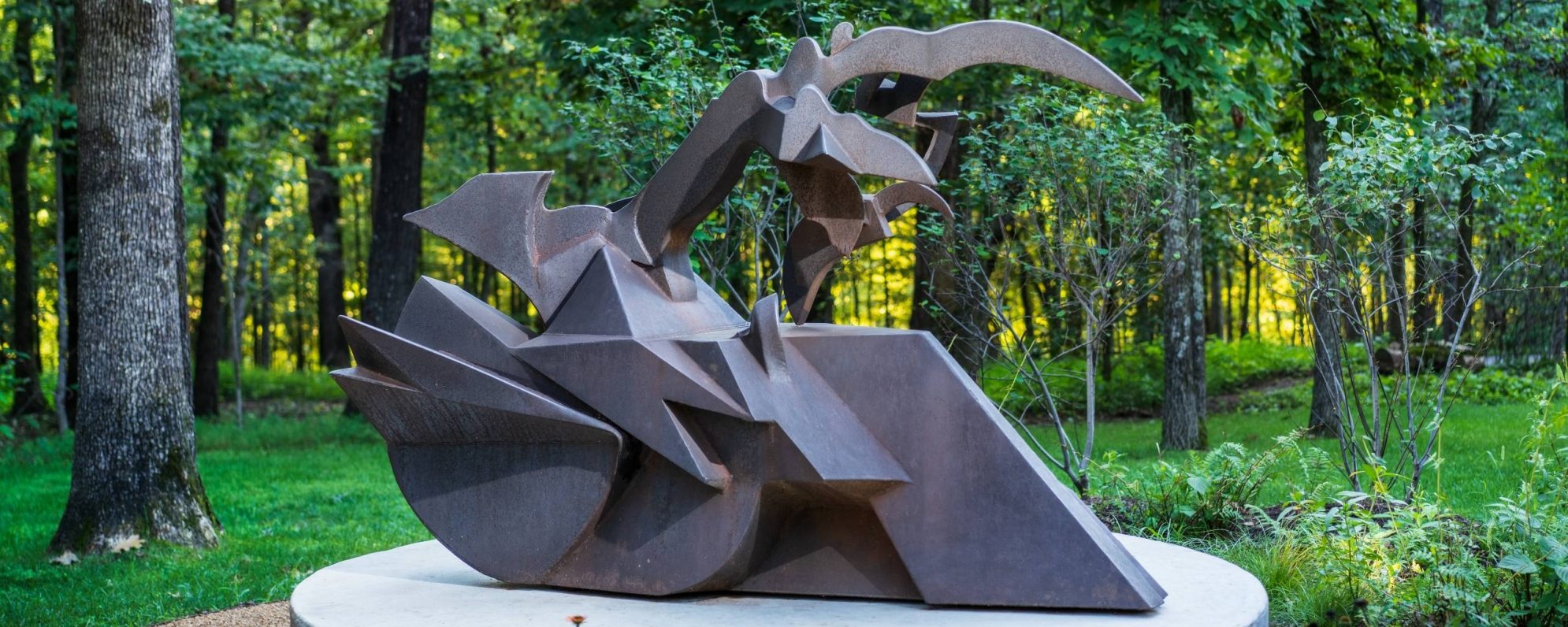 a steel gray sculpture shaped like a train engine and wheel on the bottom with a scythe shape on top sitting on a platform surrounded by a lush green forest