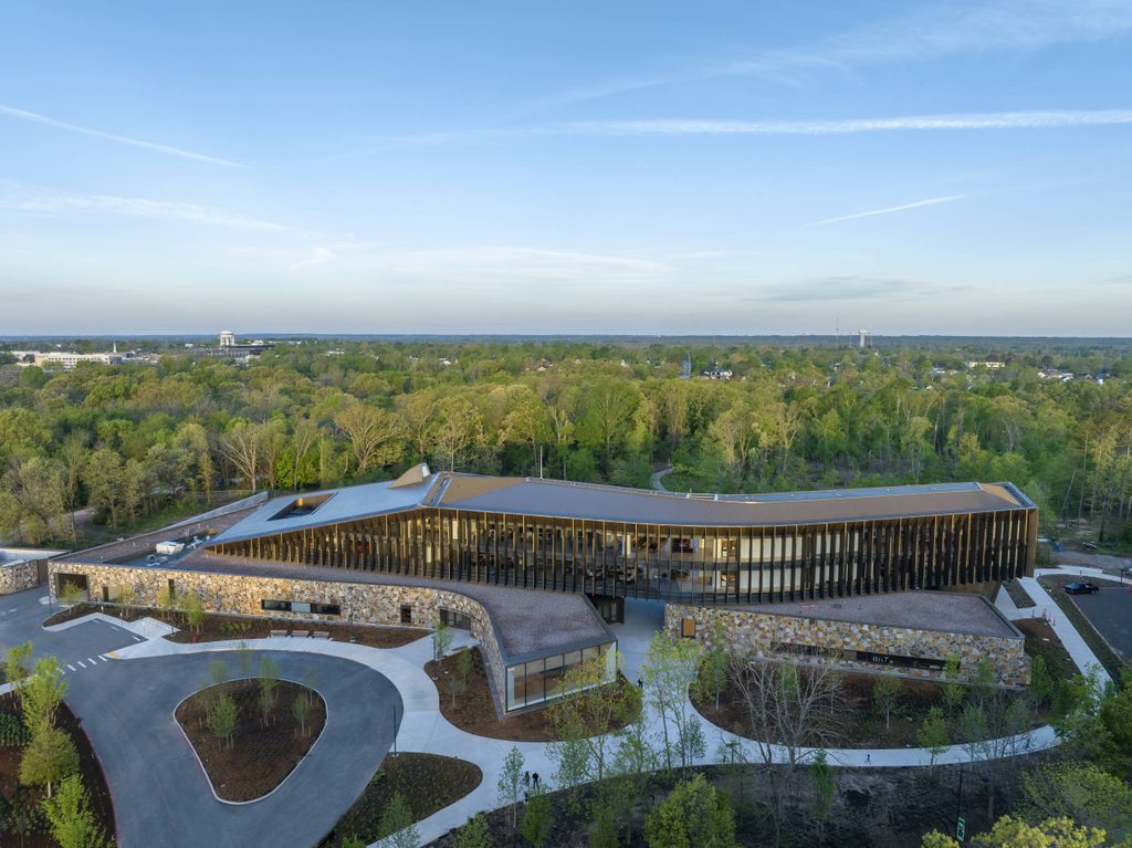 Modern large building with glass and stone, angular roof, circular driveway, surrounded by trees.