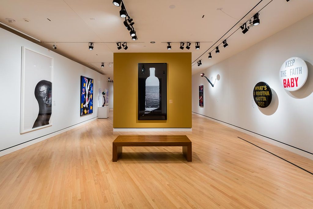 Modern art gallery with diverse artworks, framed images, text pieces, and a wooden bench.