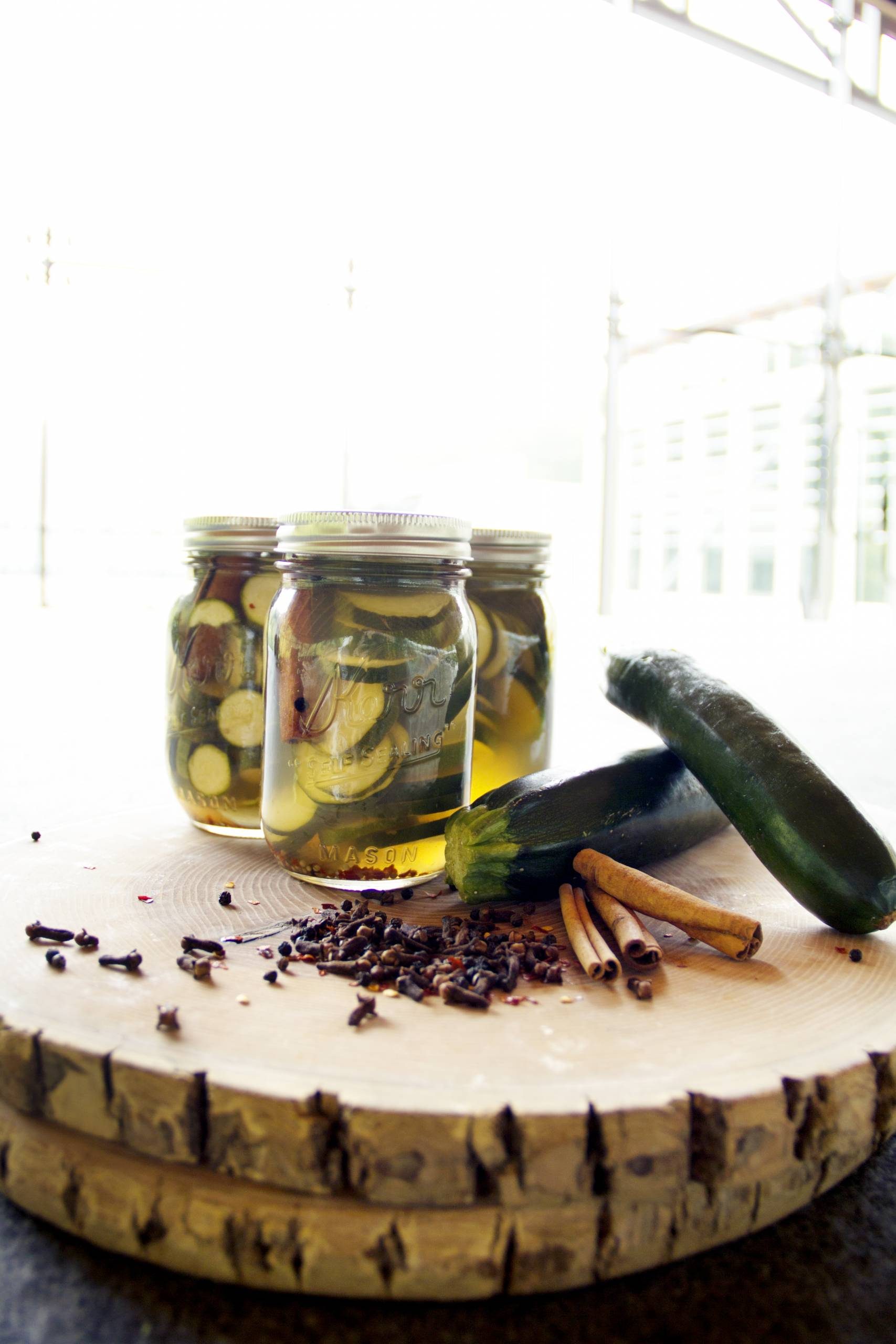 Jars of preserved vegetables on wood with fresh zucchinis, cinnamon sticks, and cloves.