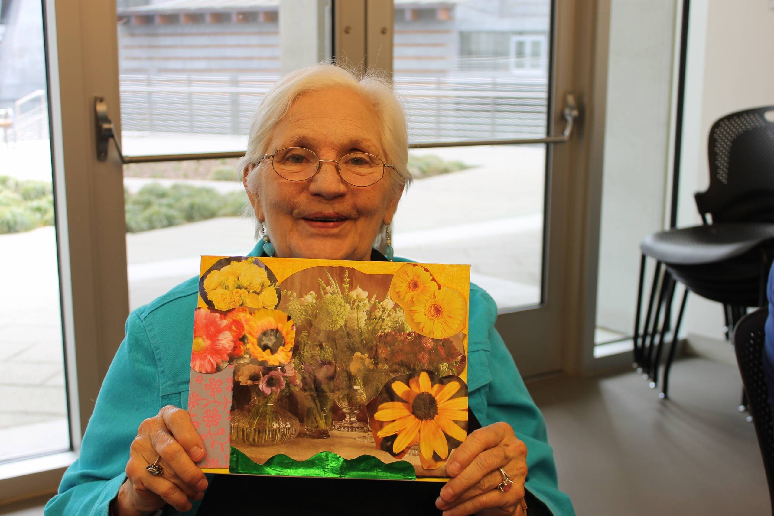Older woman in a turquoise shirt holding a colorful flower collage indoors.