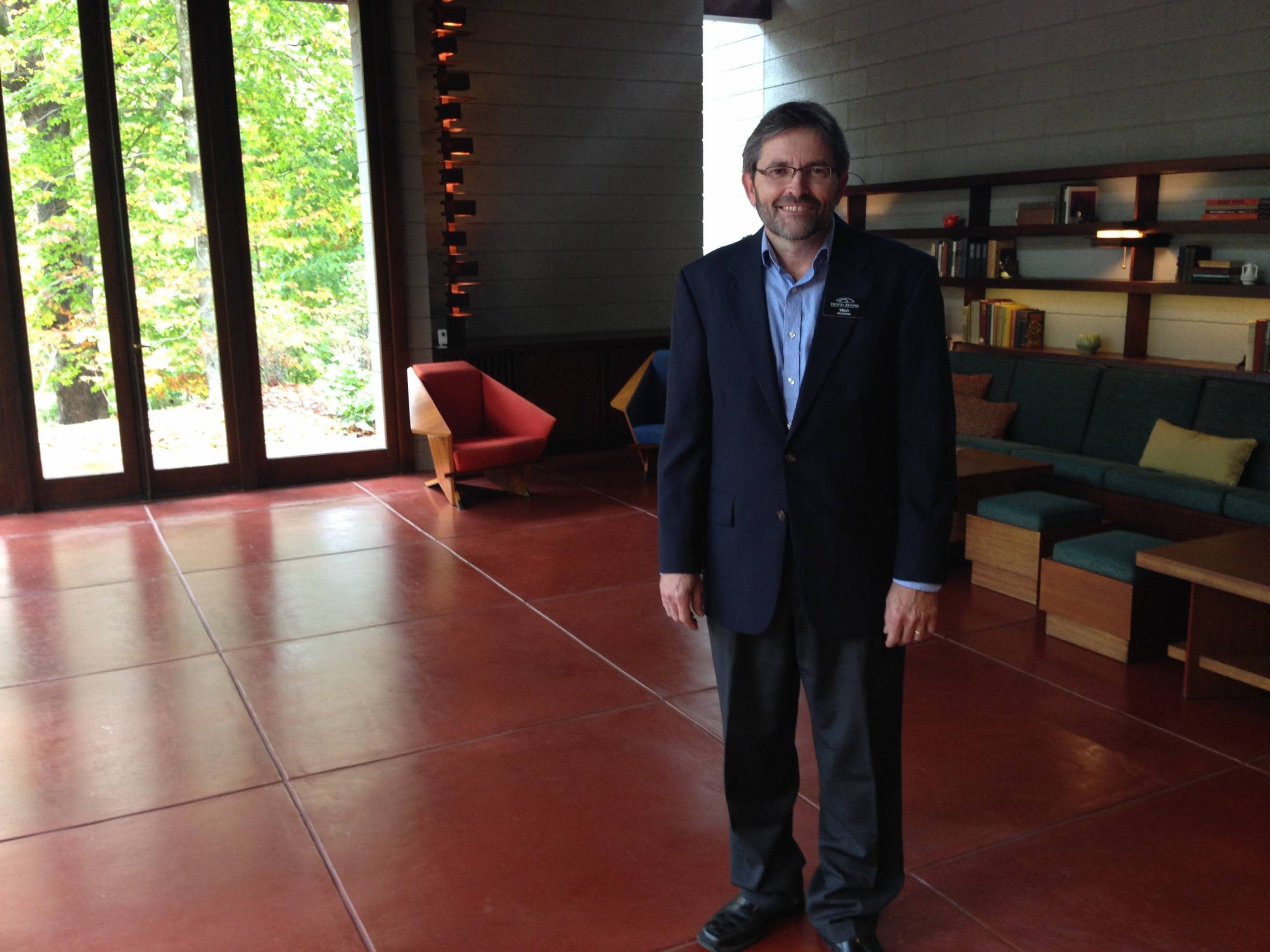 Man in a dark suit in a modern room with large windows, red floor, and contemporary furniture.