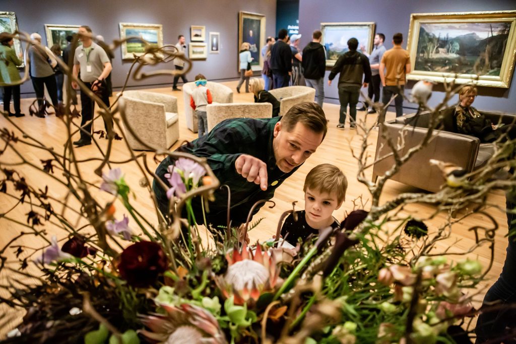 Man and child observing floral arrangement in crowded art gallery with paintings on walls.