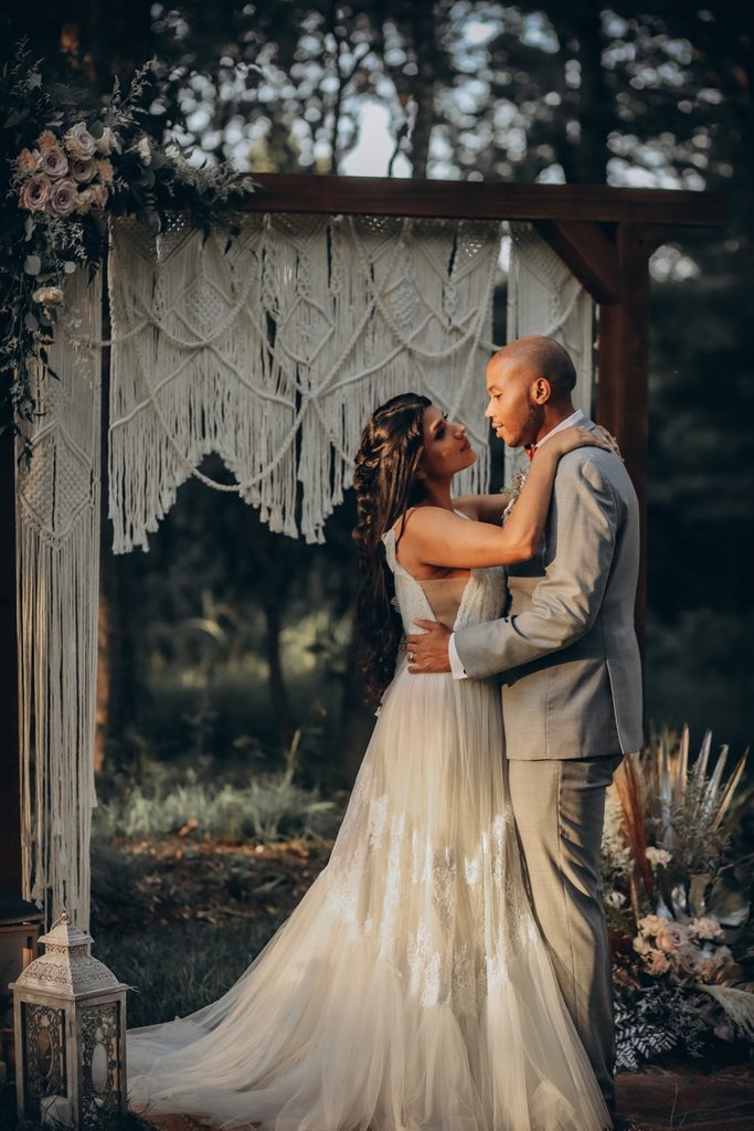 Bride and groom embrace under floral arch with macramé outdoors.