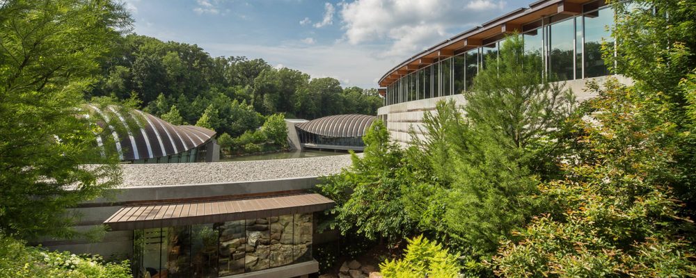 Modern building complex with glass windows, surrounded by green trees and rocky terrain under a partly cloudy sky.
