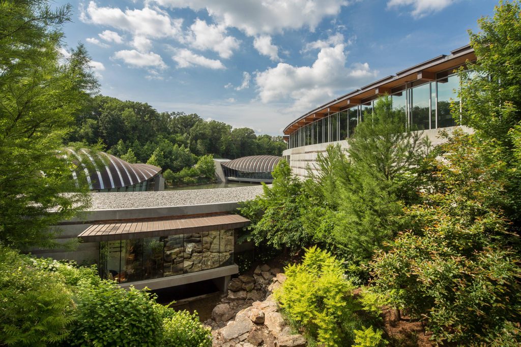 Modern building complex with glass windows, surrounded by green trees and rocky terrain under a partly cloudy sky.