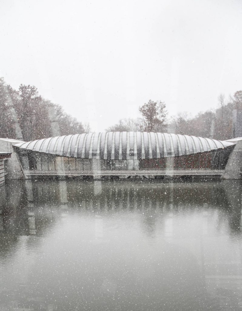 Modern building with curved roof by calm water, snow falling, trees in background.