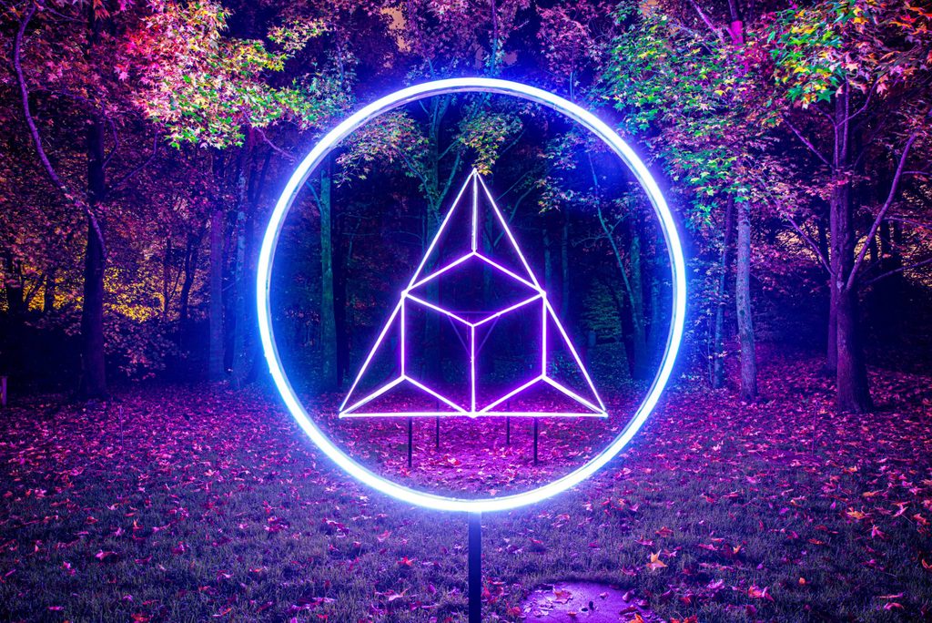 A sculpture with a cube, triangle, and circle, lit up in neon blue in the middle of a forest