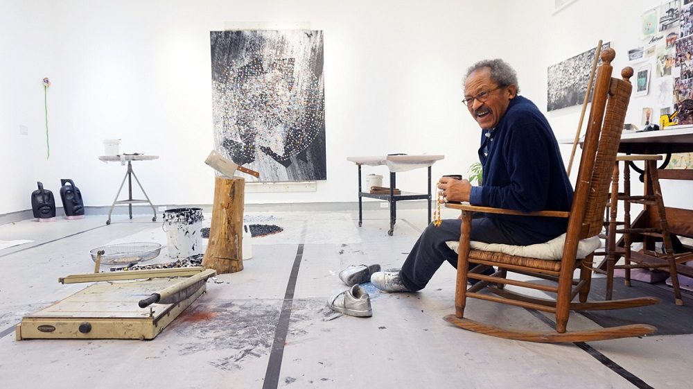 Artist in rocking chair in studio with art supplies, tools, and abstract painting on wall.