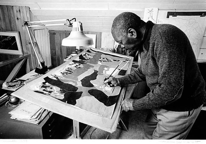 Man painting details on a large canvas in a studio with desk lamp and art materials.