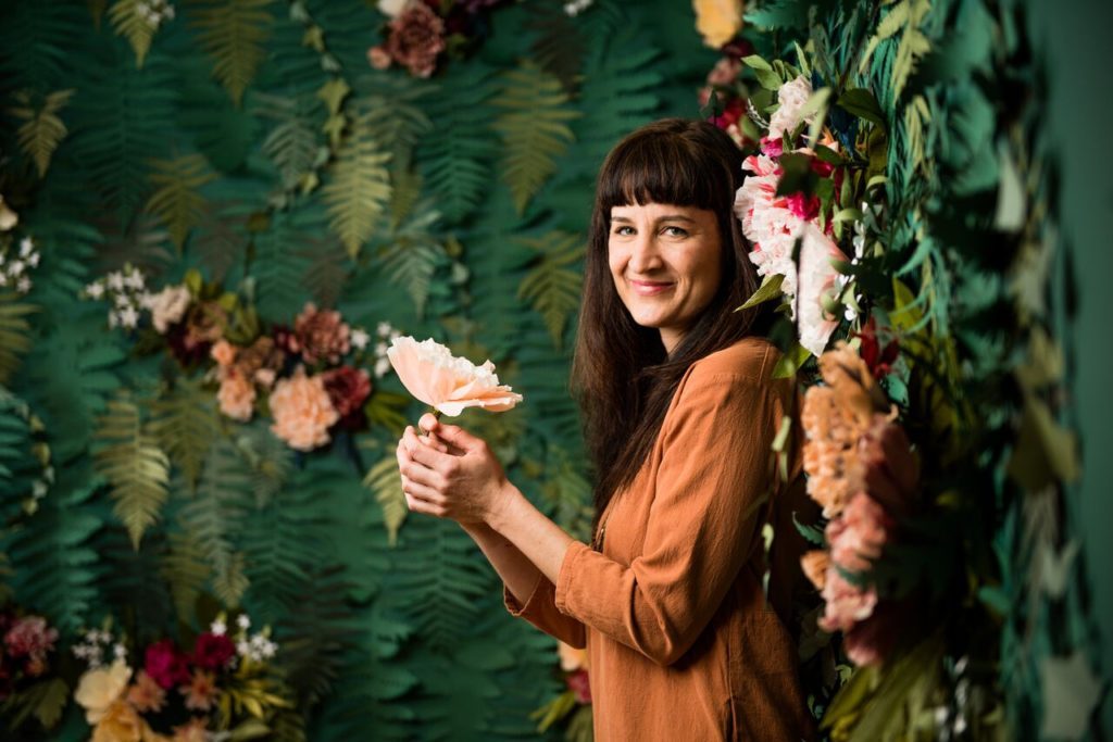 Woman with flower, in rust blouse, smiles against lush greenery and colorful blossoms.