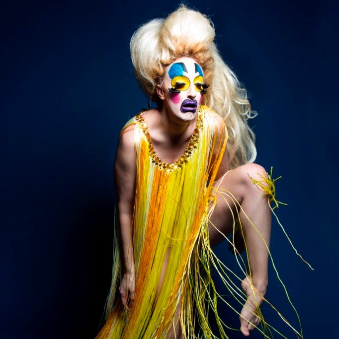 Individual in bright yellow costume with colorful makeup and voluminous blonde hair, dark backdrop.