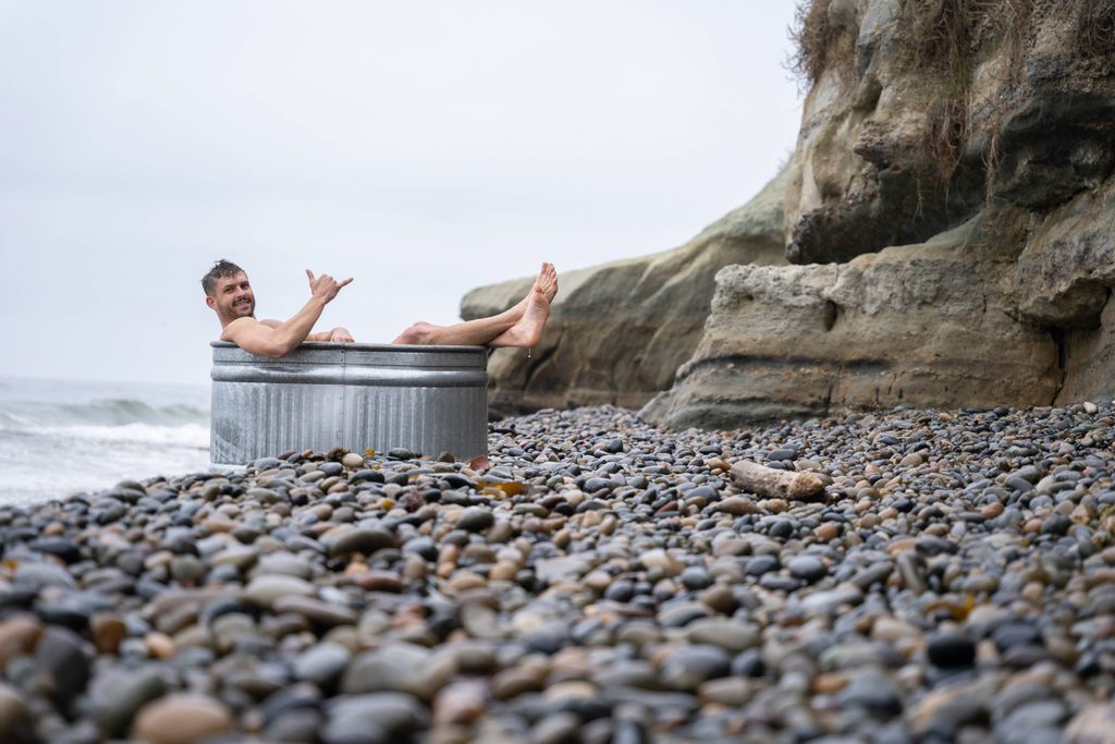 Man relaxing in a metal tub filled with water on a rocky beach near ocean cliffs.