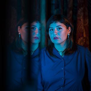 Person in dim lighting, buttoned shirt, earrings, looking at window reflection with bluish tint.