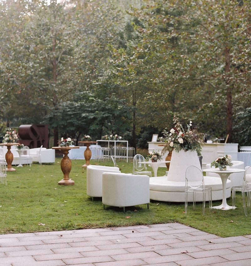 Outdoor event with white furniture, floral arrangements, on grass surrounded by trees.