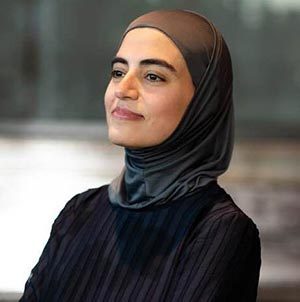 Woman in dark headscarf and outfit, side profile, blurred indoor background