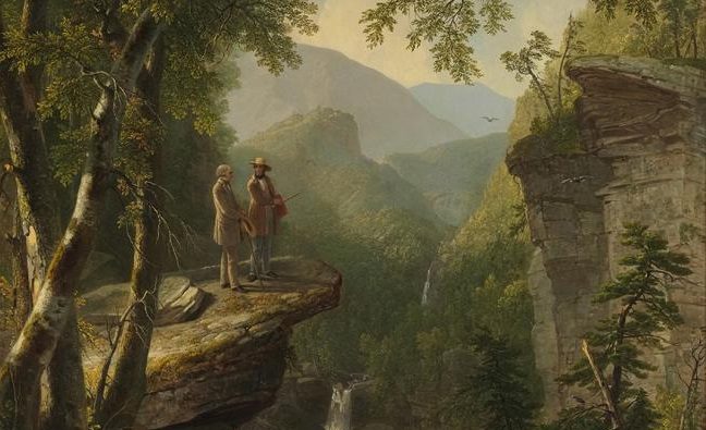 Two men stand on a rocky ledge overlooking a lush valley with a waterfall and distant mountains.