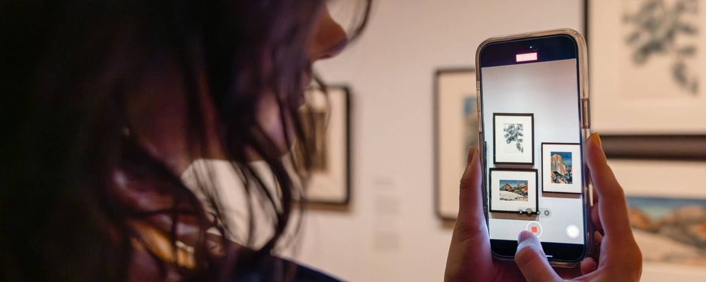 Person photographing framed artwork with a smartphone in a gallery setting.