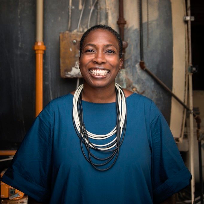 Smiling person in blue dress with necklace, industrial background with pipes and control box.